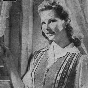 May include: A vintage black and white photograph of a woman wearing a collared top with a striped pattern. The top has a V-neck and button closure. The woman is smiling and looking towards the right.