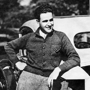 May include: A black and white photograph of a person wearing a dark gray cardigan sweater with a button-up front and two front pockets. The person is also wearing a white collared shirt and gray pants. The person is leaning against a car.