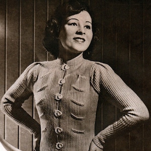 May include: A vintage, ribbed knit cardigan in a light beige colour. The long-sleeved sweater features a high collar, decorative buttons, and curved pocket details. The woman in the image is wearing the cardigan. The image is in black and white.