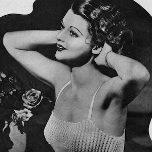 May include: A vintage black and white photograph of a woman with dark hair and dark lipstick. She is wearing a knitted top with thin straps. Her arms are raised behind her head. A vase of roses is in the background.