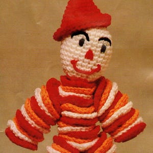 May include: A handmade, crocheted clown doll. It has a red hat and a striped outfit in red, orange, and white. The doll has a smiling face with black eyes and a red nose. The arms and legs are also striped.