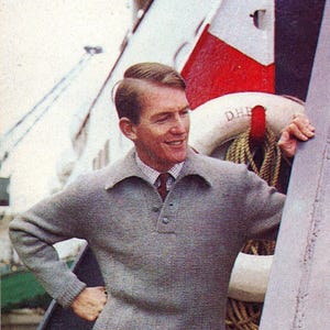 May include: A man wearing a gray knit sweater with a collared neckline and button detail. He is also wearing a patterned tie and khaki pants. The background includes a life preserver and nautical ropes.