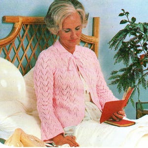 May include: A woman wearing a light pink, open-front, knitted cardigan with a tie closure. The cardigan has a delicate, openwork pattern. She is sitting on a bed and reading a red book. The headboard is made of woven bamboo.