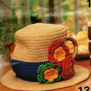 May include: A beige crocheted teapot cozy with a blue band and a matching coaster. The cozy is adorned with three colorful crocheted flowers in red, green, and orange. The teapot is on a table.