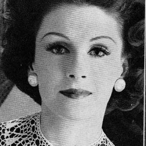 Puede incluir: Retrato en blanco y negro de una persona con cabello oscuro peinado en ondas voluminosas. La persona lleva pendientes de perlas y un cuello de encaje blanco sobre una prenda oscura. El maquillaje incluye lápiz labial oscuro y cejas definidas.