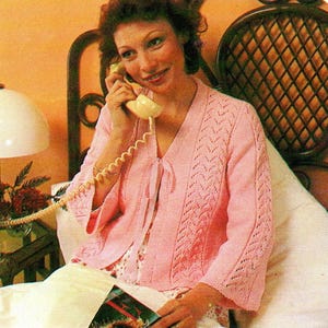 May include: A light pink, open-front cardigan with a delicate knit pattern on the sleeves and front panels. The cardigan has a tie closure at the neckline and is worn over a patterned top. The woman is holding a vintage telephone.