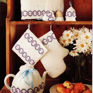 May include: A collection of white knitted kitchen items with purple and black patterned trim. Includes a tea cozy, potholders, oven mitt, and a towel. A vase of white daisies and a plate of scones are also present.