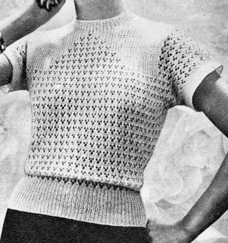 Op de afbeelding: Een off-white gebreide top met korte mouwen en een hoge halslijn. Het kledingstuk heeft een openwerkpatroon over de borst en mouwen, en een geribbelde band in de taille. De stijl is vintage, met een textuurontwerp. De mouwen zijn kort.