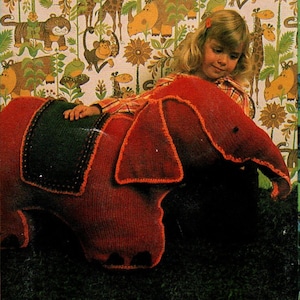 Puede incluir: Un gran cojín rojo con forma de elefante tejido, con una silla de montar verde oscuro y ribetes naranjas. El elefante tiene orejas grandes y una trompa. El fondo presenta un papel pintado colorido con temática de jungla.