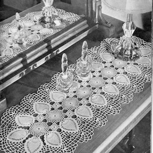 Peut inclure: Une nappe de table en crochet blanc, faite à la main, avec un motif répété de cœurs et de dentelle ajourée. La nappe est présentée sur une surface en bois foncé, avec plusieurs flacons de parfum décoratifs en verre.