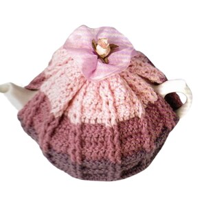 Puede incluir: Una tetera de cerámica blanca cubierta por una funda de ganchillo hecha a mano con textura acanalada en tonos degradados de rosa claro, malva y morado oscuro. La parte superior está adornada con una flor de tela rosa y un pequeño capullo.