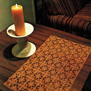 Peut inclure: Une bougie orange allumée dans un porte-bougie en céramique blanche sur une table en bois foncé. Un napperon en dentelle orange avec un motif floral est posé sur la table. Un canapé rayé est en arrière-plan.