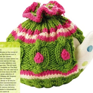 May include: A green, pink, and white knitted tea cozy. The cozy has a ruffled pink top and decorative leaf-like patterns. The teapot is white with blue and green polka dots.