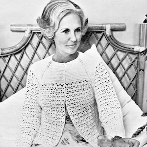 Peut inclure: Un cardigan blanc en crochet avec un col rond et des manches longues. Le motif ajouré présente un motif texturé. Le cardigan est porté sur un haut blanc. L'arrière-plan comprend une tête de lit en osier et un verre d'eau.