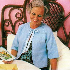 Op de afbeelding: Een lichtblauwe gebreide cardigan met korte mouwen en een striksluiting. De cardigan wordt gedragen over een top met bloemenprint. De vrouw houdt een wenskaart met bloemenillustraties vast. De achtergrond omvat een rotan hoofdbord en een roze muur.