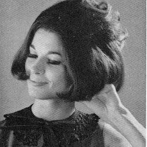 May include: Black and white image of a woman with a bouffant hairstyle. The hair is styled with volume at the crown and sides, framing the face. She is wearing a dark top with a detailed neckline.