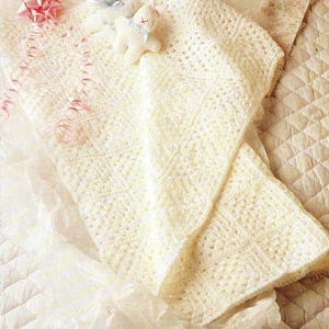 Könnte beinhalten: Cremefarbene, gehäkelte Babydecke mit strukturiertem Muster. Die Decke ist gefaltet und über Seidenpapier drapiert, mit einer rosa Schleife und zwei kleinen Stofftieren mit blauen Bändern. Eine weiche, zarte Ästhetik.