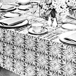May include: A black and white image of a formal dining table setting. The table is covered with a white, intricately crocheted tablecloth. White plates, silverware, and glassware are set for each place setting. A floral arrangement and salt and pepper shakers are also visible.