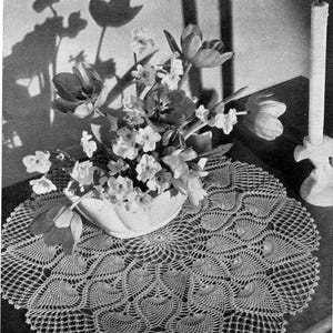 Puede incluir: Un tapete de ganchillo con un diseño calado detallado, con un arreglo floral en un cuenco blanco. El arreglo incluye tulipanes y otras flores. Una vela blanca y su soporte están a la derecha.