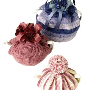 Könnte beinhalten: Drei gestrickte Teewärmer. Einer ist blau-violett gestreift mit blauem Band und Stoffblume. Einer ist einfarbig malvenfarben mit dunkelroter Stoffblume. Einer ist cremefarben-malvenfarben gestreift mit passendem Bommel. Alle haben sichtbare Ausgüsse.