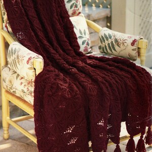 May include: A deep burgundy knitted throw, featuring a detailed pattern and tassels, draped over a chair with floral upholstery. The blanket's rich colour and texture create a cosy feel. The tassels provide a decorative finish.