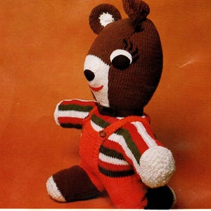 Puede incluir: Un juguete de oso tejido a mano con cabeza marrón, cara blanca y peto rojo. El oso tiene ojos negros, una sonrisa roja y mangas a rayas. El juguete está sobre un fondo naranja.