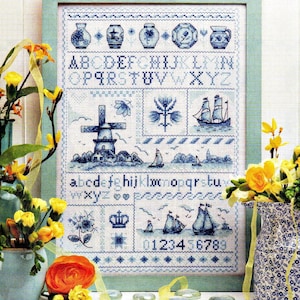 以下が含まれることがあります： 白い布に青い糸で刺繍されたクロスステッチのサンプラー。大文字と小文字のアルファベット、0から9までの数字、風車、帆船、花のモチーフ、王冠、様々な陶器の形がグリッド状に配置され、額縁に入れられている。