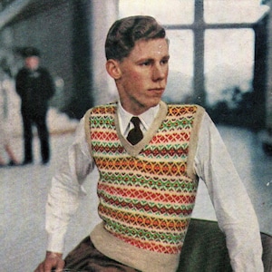 Op de afbeelding: Een mouwloos V-hals spencer vest met een herhalend geometrisch patroon in tinten rood, groen, geel en wit. Het kledingstuk heeft een effen beige geribbelde boord langs de halslijn, armsgaten en zoom, gedragen over een wit overhemd met kraag.