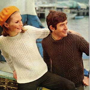 May include: A vintage photograph featuring two people wearing knitted sweaters. One person wears a cream-colored sweater and an orange beret. The other person wears a brown sweater. The background shows a waterfront scene with boats.