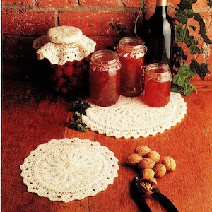 Op de afbeelding: Een rustig stilleven met potten jam, een fles wijn en gehaakte kleedjes. De potten bevatten een roodbruine jam en zijn bedekt met kant. Walnoten en een notenkraker zijn ook aanwezig.