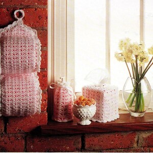 May include: A collection of handmade crochet items in a bathroom setting. Includes a pink and white crochet toilet paper holder, a tissue box cover, a small bag, and a decorative bowl. A vase of flowers sits on the windowsill.