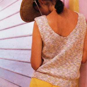 May include: A straw sun hat with a wide brim and a sleeveless knit top in beige and yellow hues. The model is wearing yellow shorts. The background features a pink and white wall.