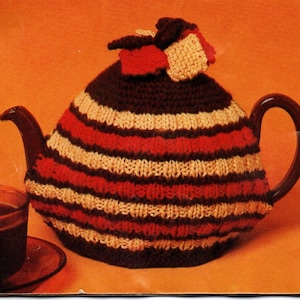 Könnte beinhalten: Eine braune Teekanne mit einem gestrickten Teewärmer. Der Teewärmer hat horizontale Streifen in Rot, Creme und Dunkelbraun. Eine passende Strickverzierung sitzt obenauf. Eine Tasse Tee ist im Vordergrund zu sehen.