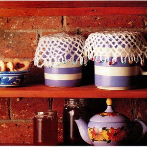 May include: A cozy kitchen scene with a wooden shelf holding a blue and white bowl of heart-shaped cookies, two mugs with crocheted covers, and a floral teapot. The mugs are purple with white stripes.