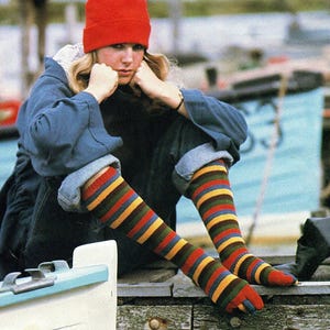 May include: A person wearing a red knit cap, blue jacket, and striped knee-high socks with red, yellow, green, and blue stripes. The person is sitting on a wooden dock near a boat.