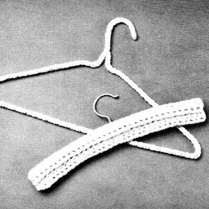 May include: A white, crocheted clothing hanger with a matching crocheted sleeve. The hanger is made of a thick, textured yarn. The sleeve is designed to fit over the hanger's bar, adding a decorative touch. The background is a solid gray.