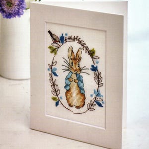May include: A white greeting card with a cross-stitch design featuring a brown rabbit wearing a blue jacket, surrounded by a floral wreath. A small bird is perched on the wreath. The card is displayed at an angle.