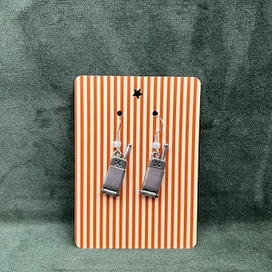 May include: Silver-toned walkie-talkie earrings hanging from a striped orange and white display card. The earrings have a detailed design and dangle from silver hooks. The card has a star cutout at the top.