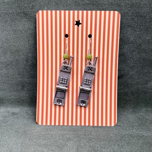 May include: A pair of silver-toned earrings shaped like vintage cell phones. Each earring has a green bead and a small hook. The earrings are displayed on a red and white striped card with a black star.