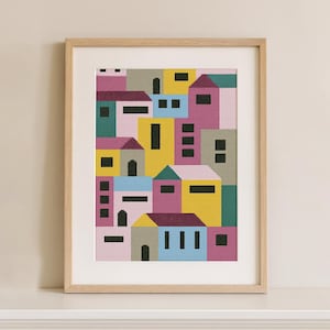 Puede incluir: Lámina artística enmarcada que representa un paisaje urbano abstracto. La obra presenta un mosaico de casas geométricas en tonos rosa, amarillo, verde azulado y azul, con ventanas y puertas negras. La impresión está enmarcada en un marco de madera natural.