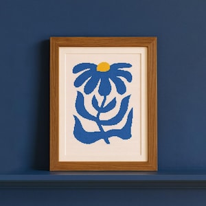 May include: A framed artwork with a blue flower design and a yellow centre. The artwork is in a wooden frame and is displayed against a blue background. The flower has a stylized, geometric appearance.