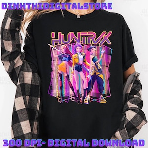 Può includere: T-shirt nera con una grafica di tre figure animate in abiti colorati. La parola "HUNTRAX" è visualizzata in un carattere vibrante e stilizzato sopra le figure. La frase "300 DPI - DOWNLOAD DIGITALE" è stampata in basso.