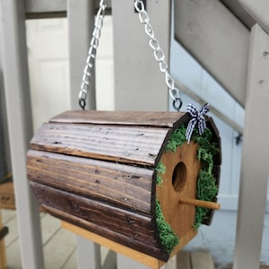 May include: A rustic, wooden birdhouse with a dark brown finish, hanging from a silver chain. The birdhouse has a round entrance, a wooden perch, and is decorated with green moss and a black and white checkered bow.