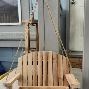 May include: A wooden hanging bird feeder with a slatted back and armrests. The feeder is suspended by rope and attached to a black metal bracket. The natural wood color contrasts with the light blue and gray background.