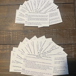 Puede incluir: Dos conjuntos en forma de abanico de tarjetas blancas con texto negro, sobre una superficie de madera. Las tarjetas contienen texto religioso, como "Taking His Name upon Me" y "Becoming Better."