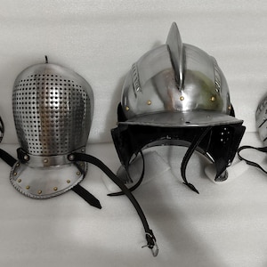 May include: Four pieces of medieval armor, including helmets and face guards, are displayed. The metal pieces are silver-colored, with leather straps and rivets. The helmets vary in design, showcasing historical craftsmanship.