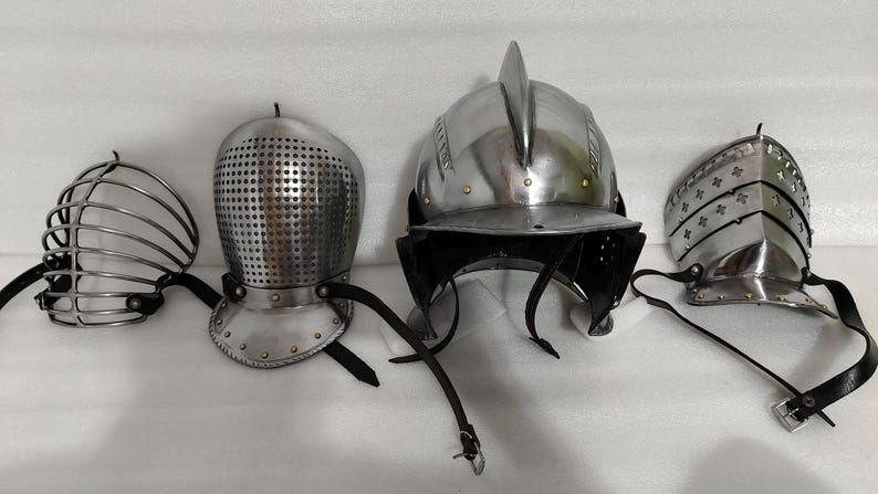 May include: Four pieces of medieval armor, including helmets and face guards, are displayed. The metal pieces are silver-colored, with leather straps and rivets. The helmets vary in design, showcasing historical craftsmanship.