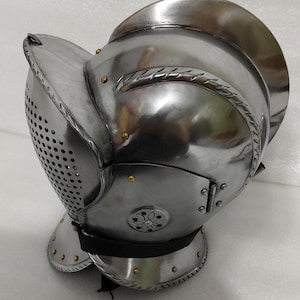 May include: A polished steel helmet with a visor featuring numerous small holes for ventilation. The helmet has a rounded top and a hinged face guard, with decorative rivets and edging. It is likely a piece of historical armor.