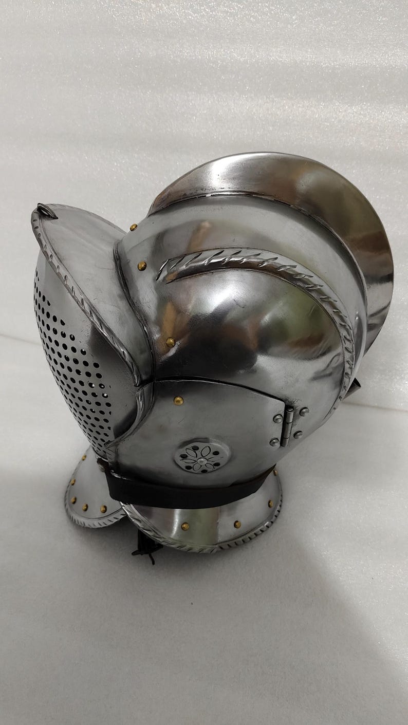 May include: A polished steel helmet with a visor featuring numerous small holes for ventilation. The helmet has a rounded top and a hinged face guard, with decorative rivets and edging. It is likely a piece of historical armor.
