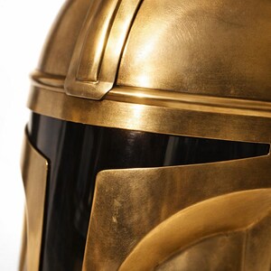 May include: Close-up of a gold-colored metal helmet with a black visor. The helmet has a detailed design with visible seams and edges. The text "Handmade Metal Construction" is displayed on the lower portion of the image.